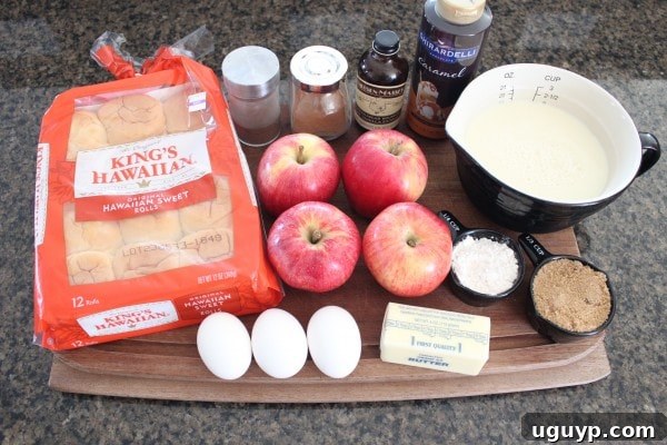 Various ingredients for apple bread pudding laid out on a table, including Hawaiian rolls, apples, eggs, milk, and spices.