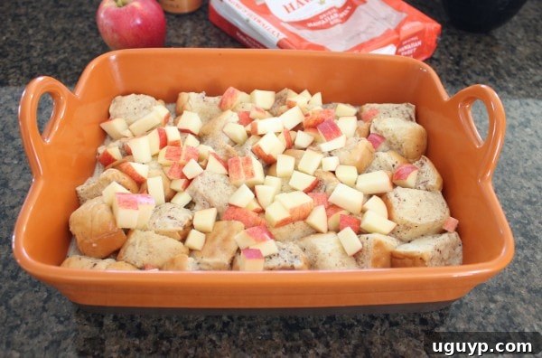 Diced apples spread over the unbaked bread pudding mixture in an orange baking dish, ready to be stirred in.