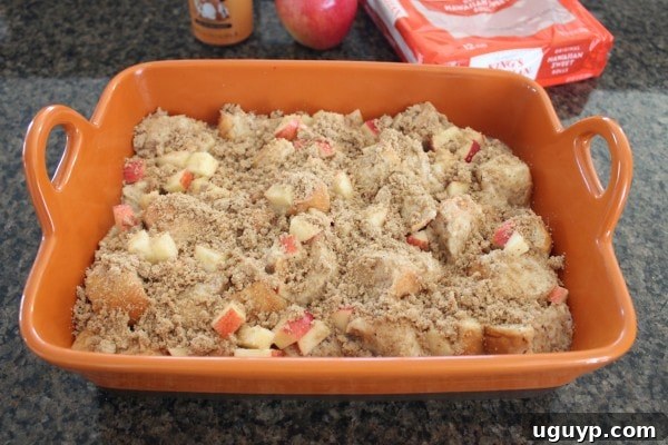Unbaked bread pudding in a baking dish, generously topped with diced apples and a golden-brown sugar crumble, ready for the oven.
