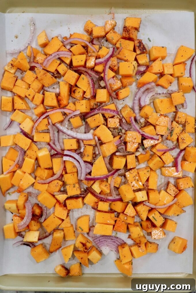 Cubed butternut squash, thinly sliced red onions, and minced garlic tossed with olive oil and spices, spread on a parchment-lined baking sheet, ready for roasting.