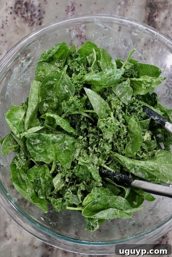 Fresh baby spinach and chopped kale being gently tossed with a creamy green dressing in a large glass mixing bowl, ready for salad assembly.