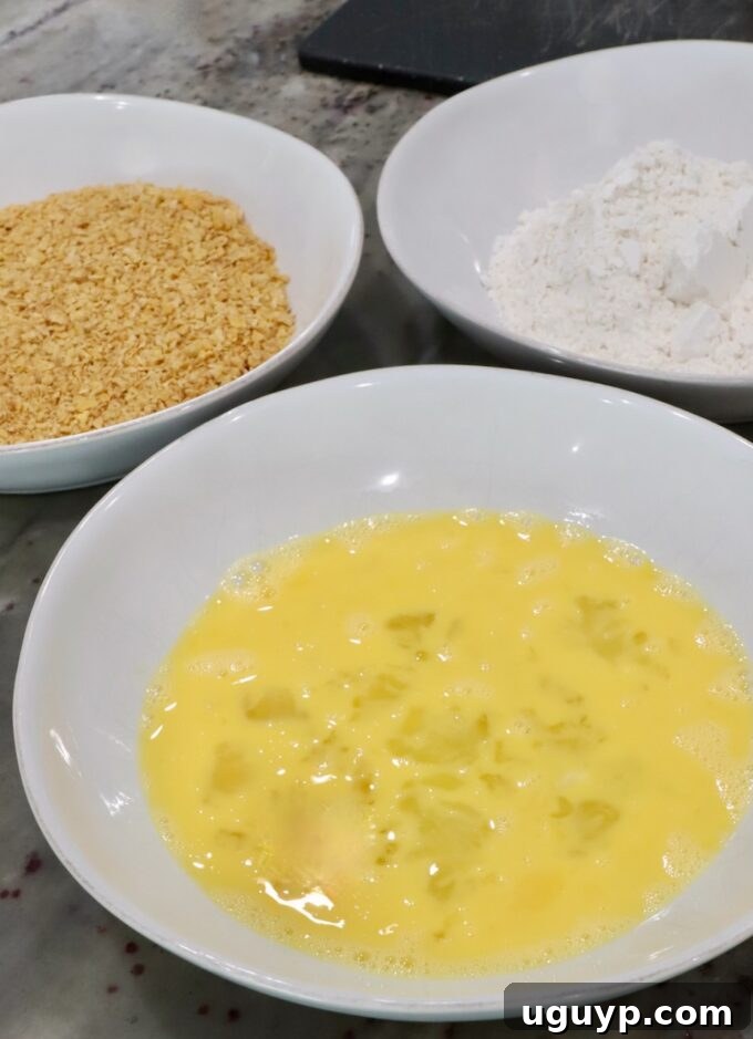 Three bowls on a countertop: one with whisked eggs, one with flour, and one with crushed cornflakes