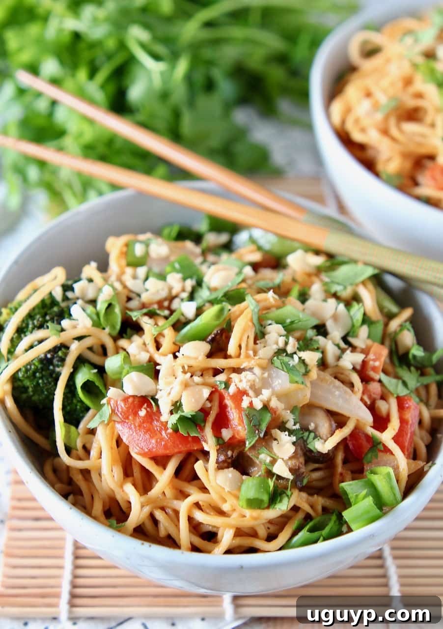 A close-up view of flavorful ramen noodles and crisp stir-fried vegetables generously coated in a rich Thai peanut sauce, presented in a ceramic bowl with chopsticks resting beside it.