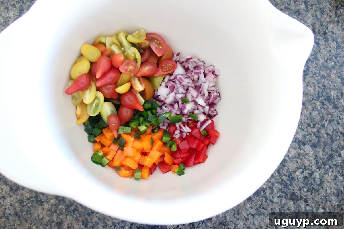 A vibrant collection of freshly diced red bell pepper, red onion, and jalapeño, alongside halved cherry tomatoes, all gathered in a spacious mixing bowl, eagerly awaiting the cooked Israeli couscous.