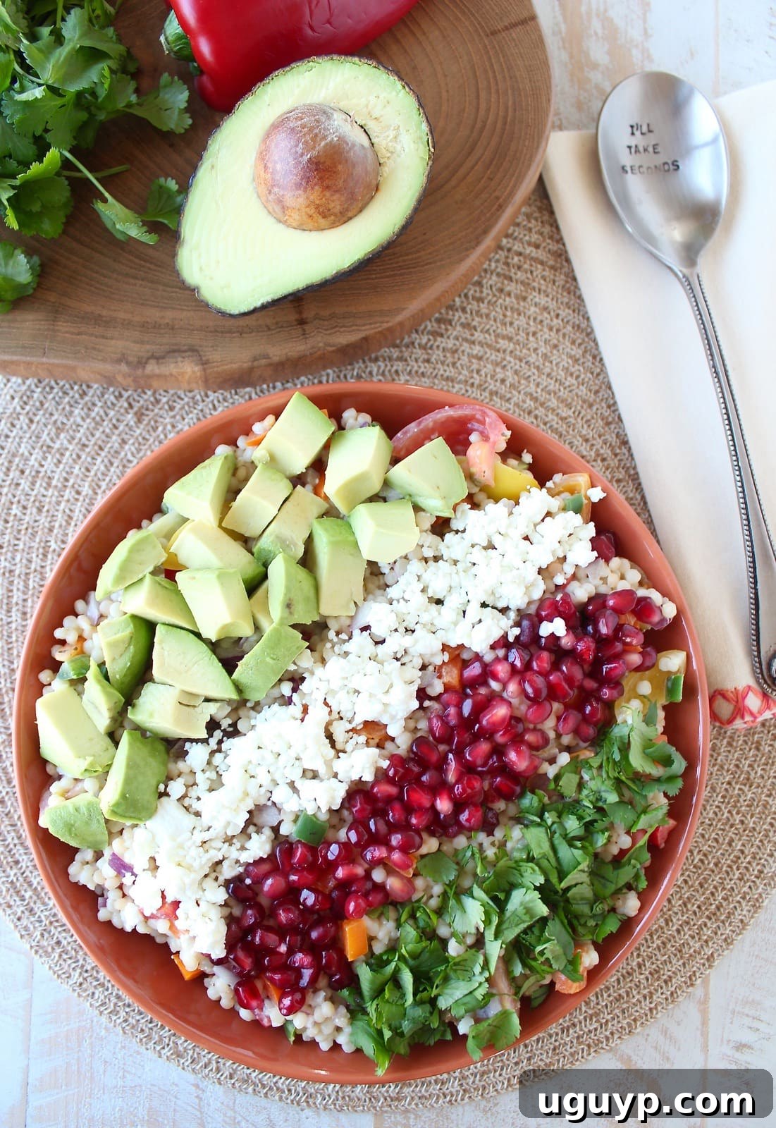 The finished Israeli Couscous Avocado Salad, beautifully arranged in an orange bowl, artfully garnished with fresh cilantro and vibrant pomegranate seeds, showcasing its enticing appeal.