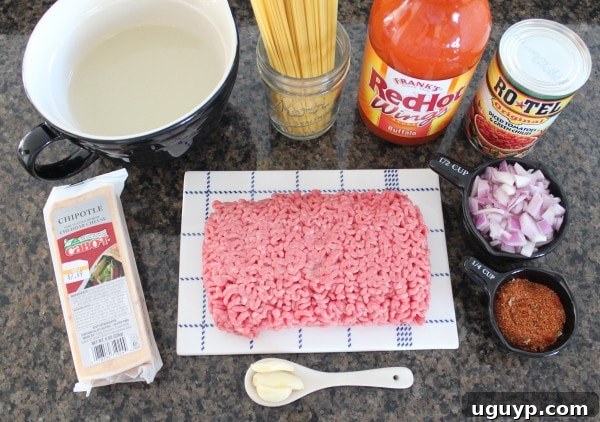 A selection of fresh ingredients including red onion, garlic, ground beef, spaghetti, Rotel, and buffalo sauce laid out on a white surface, ready for cooking.