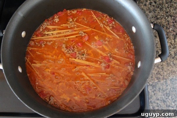 Noodles, beef, diced tomatoes, and chicken broth simmering together in a large pot, the pasta absorbing the liquid.