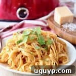 Crock pot chicken alfredo on white plate, with red crock pot in background sitting next to wood cutting board with parmesan cheese