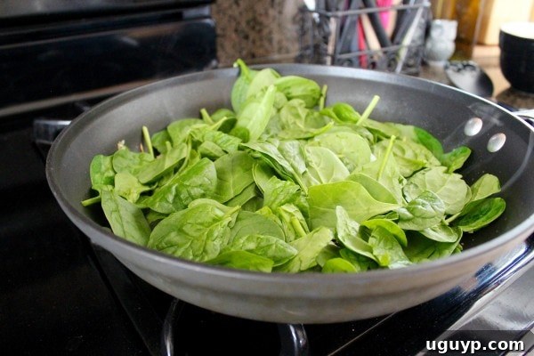 Fresh spinach wilting in a large skillet with diced onions and minced garlic