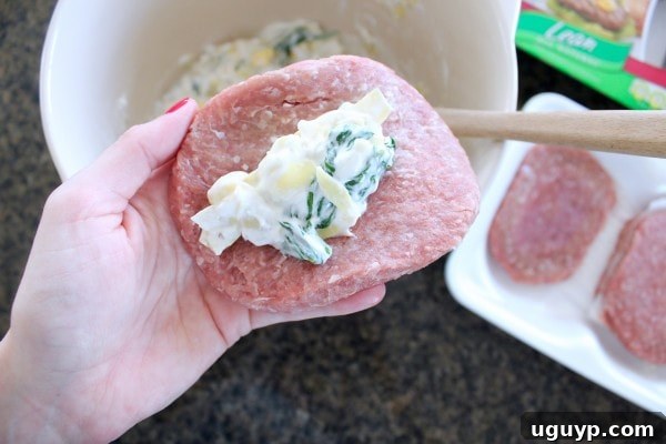 A hand carefully shaping a turkey burger patty around a spoonful of spinach artichoke dip