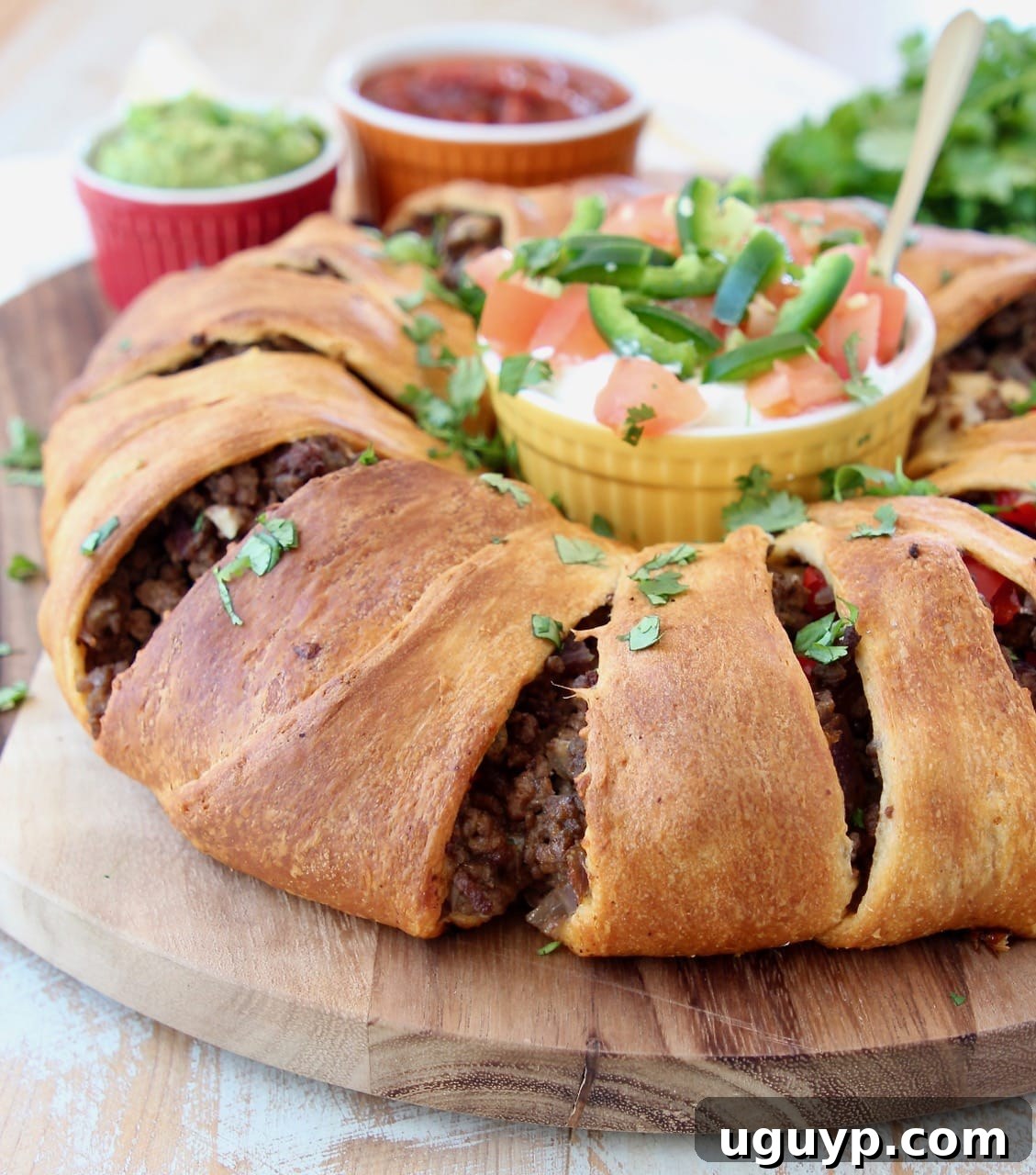 A beautifully baked taco ring made with golden crescent rolls, filled with seasoned ground beef, peppers, and onions, garnished with fresh tomatoes and jalapeños, served on a cutting board with a ramekin of sour cream.