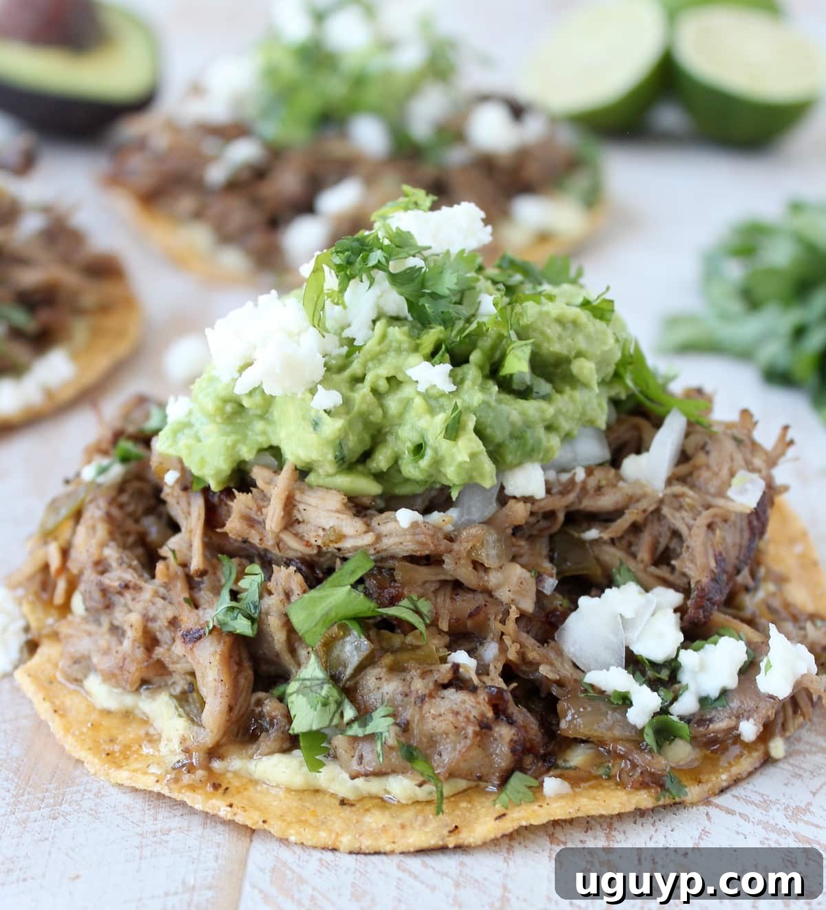 A close-up of tender, shredded pork on a crispy tostada, beautifully garnished with fresh guacamole and crumbled cheese, ready for a delicious bite.
