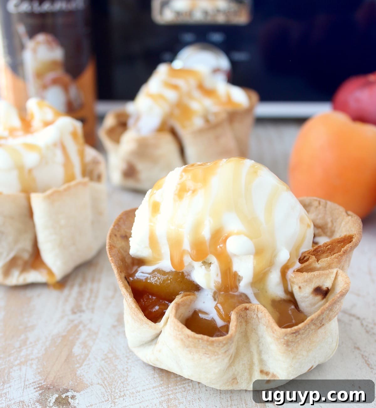 crispy taco shell cups filled with slow cooked fruit, topped with a scoop of ice cream and caramel syrup on a white wood board in front of a blue crock pot