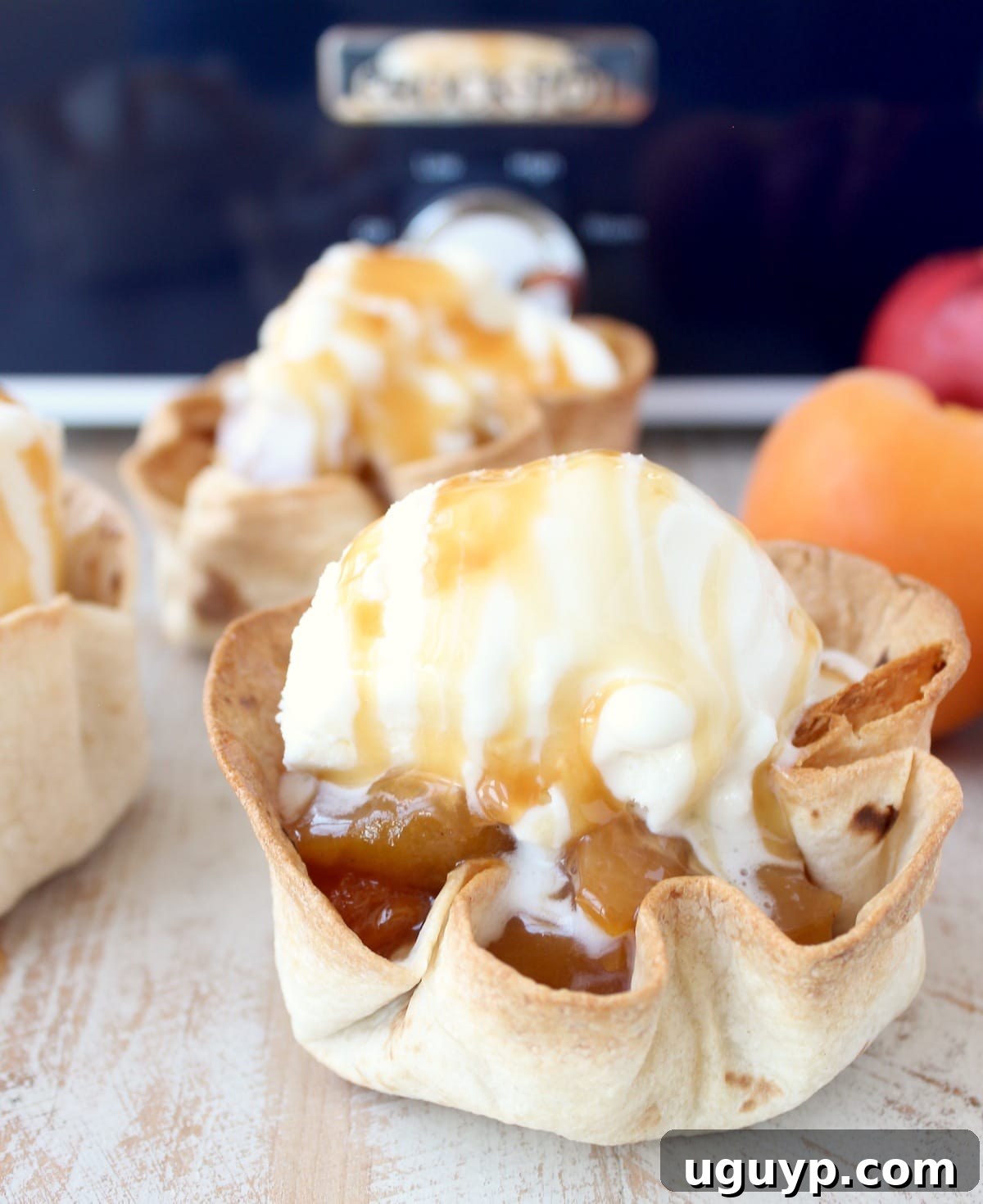 crispy taco shell cups filled with slow cooked fruit, topped with a scoop of ice cream and caramel syrup on a white wood board in front of a blue crock pot