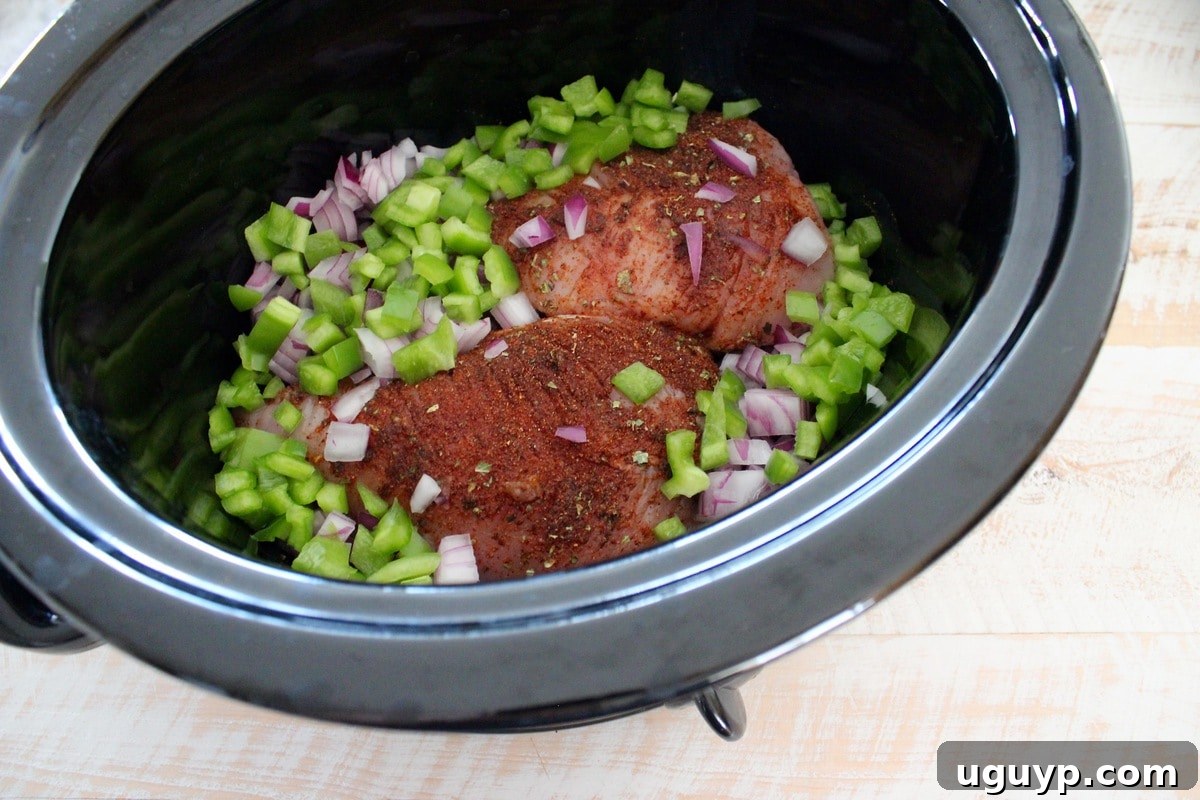 A black crock pot bowl filled with seasoned chicken, diced peppers, and onions, ready for the next ingredients.