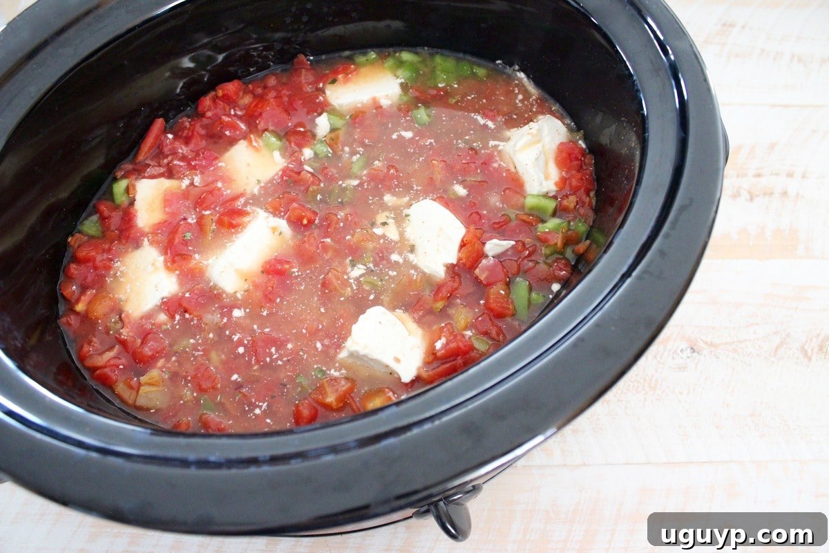 A slow cooker filled with diced peppers, diced tomatoes with green chilies, chicken broth, and chunks of cream cheese, ready to be cooked.