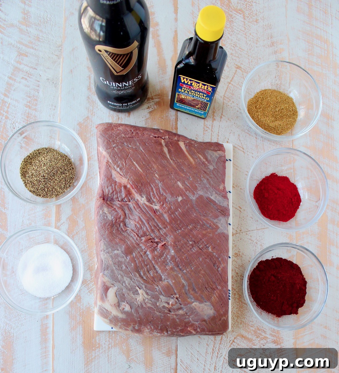 A collection of ingredients for making brisket tacos laid out on a white wooden board, including a slab of brisket, spices, and a bottle of stout beer.