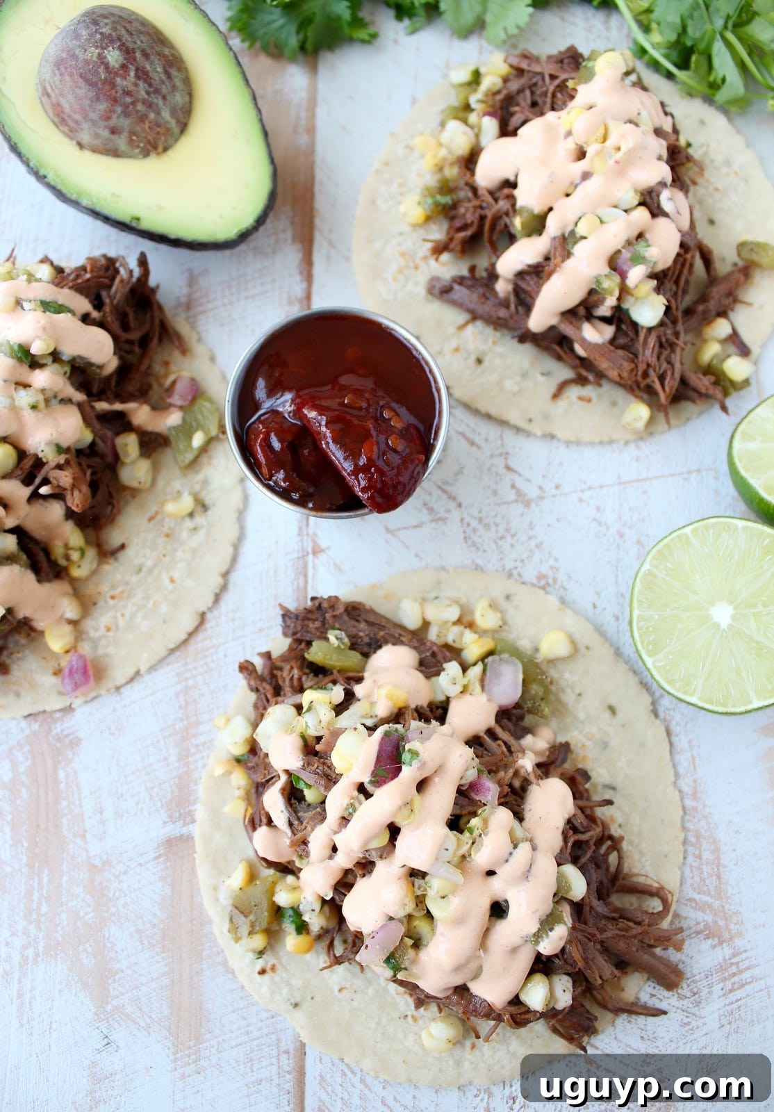 A close-up of shredded cooked brisket on corn tortillas, topped with fresh corn salsa and a generous drizzle of creamy chipotle sauce, garnished with cilantro.