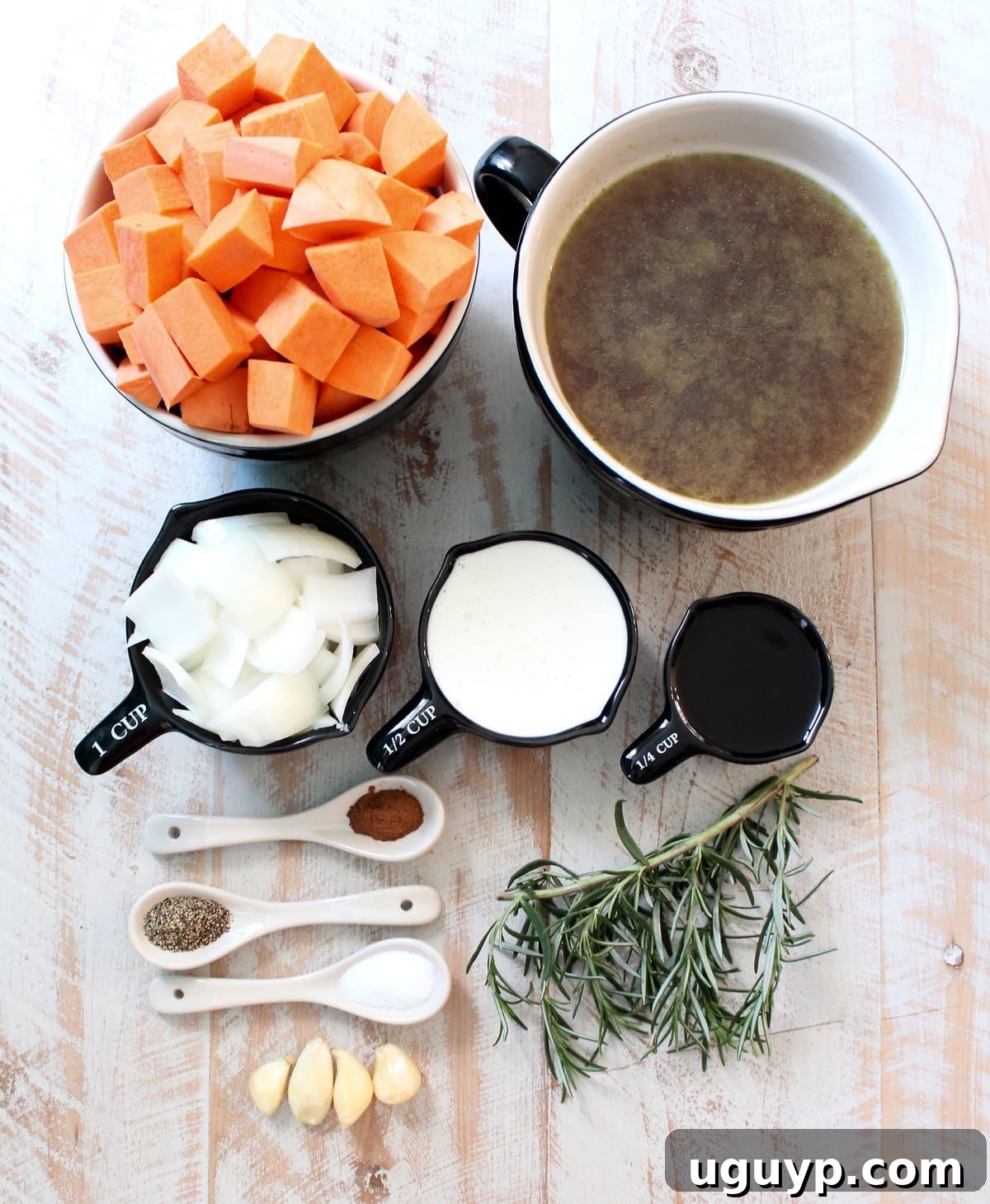 A selection of fresh ingredients for sweet potato soup laid out on a white wooden board, including sweet potatoes, onion, garlic, rosemary, and cinnamon.