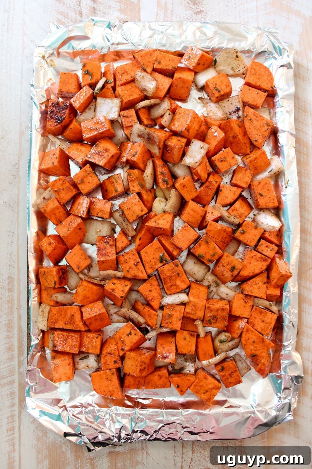 Cubed sweet potatoes, diced onion, and whole garlic cloves are spread evenly on a foil-lined baking sheet, ready for roasting.