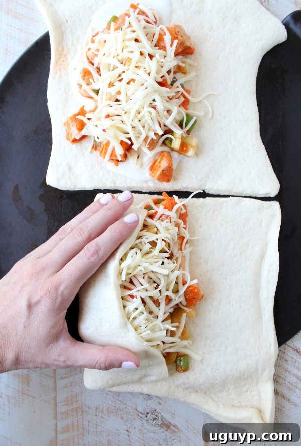 A hand gently folding uncooked pizza dough over a generous filling of buffalo chicken, celery, carrots, green onions, and melted cheese, preparing it for baking.