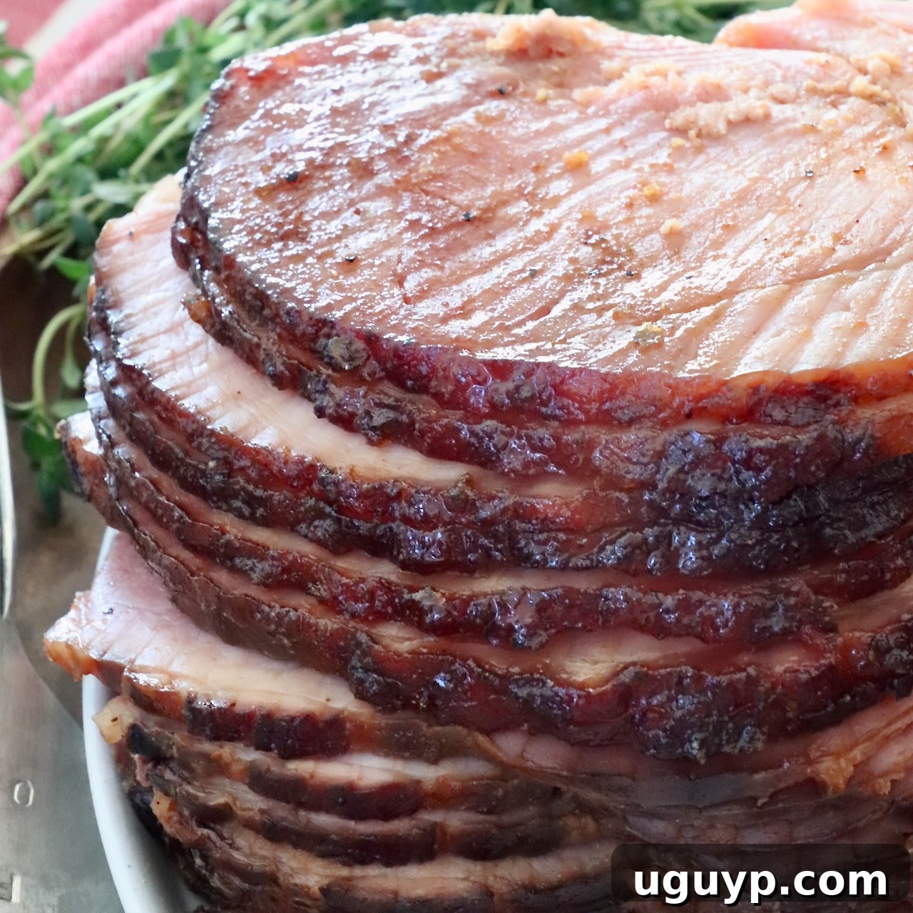 Sliced spiral ham glazed to a shiny brown, stacked on a white plate. Fresh herbs in the background and a red-striped cloth