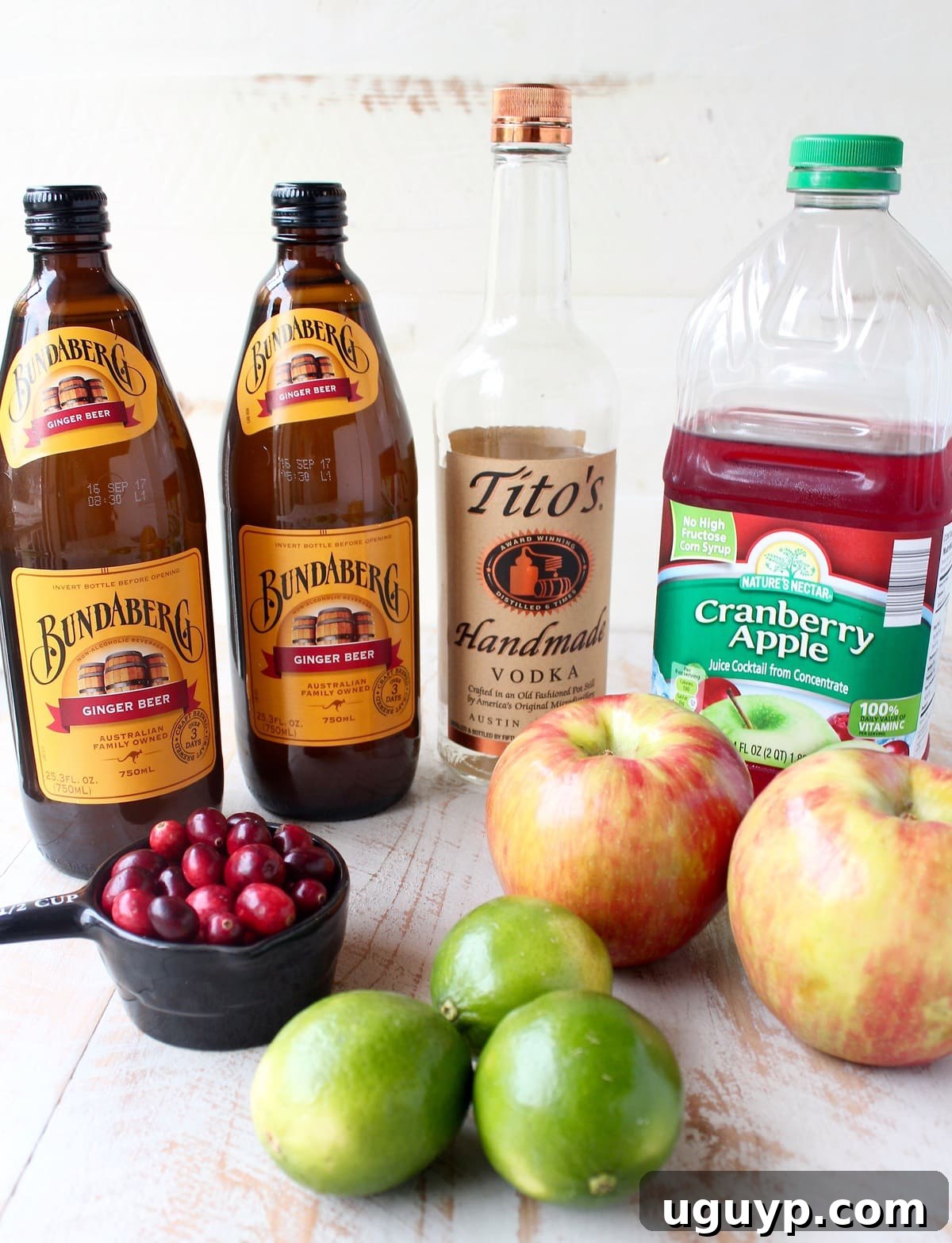 Ingredients for cranberry apple Moscow Mule punch laid out on a white wooden board, showcasing fresh cranberries, diced apples, limes, ginger beer bottles, and a bottle of vodka.