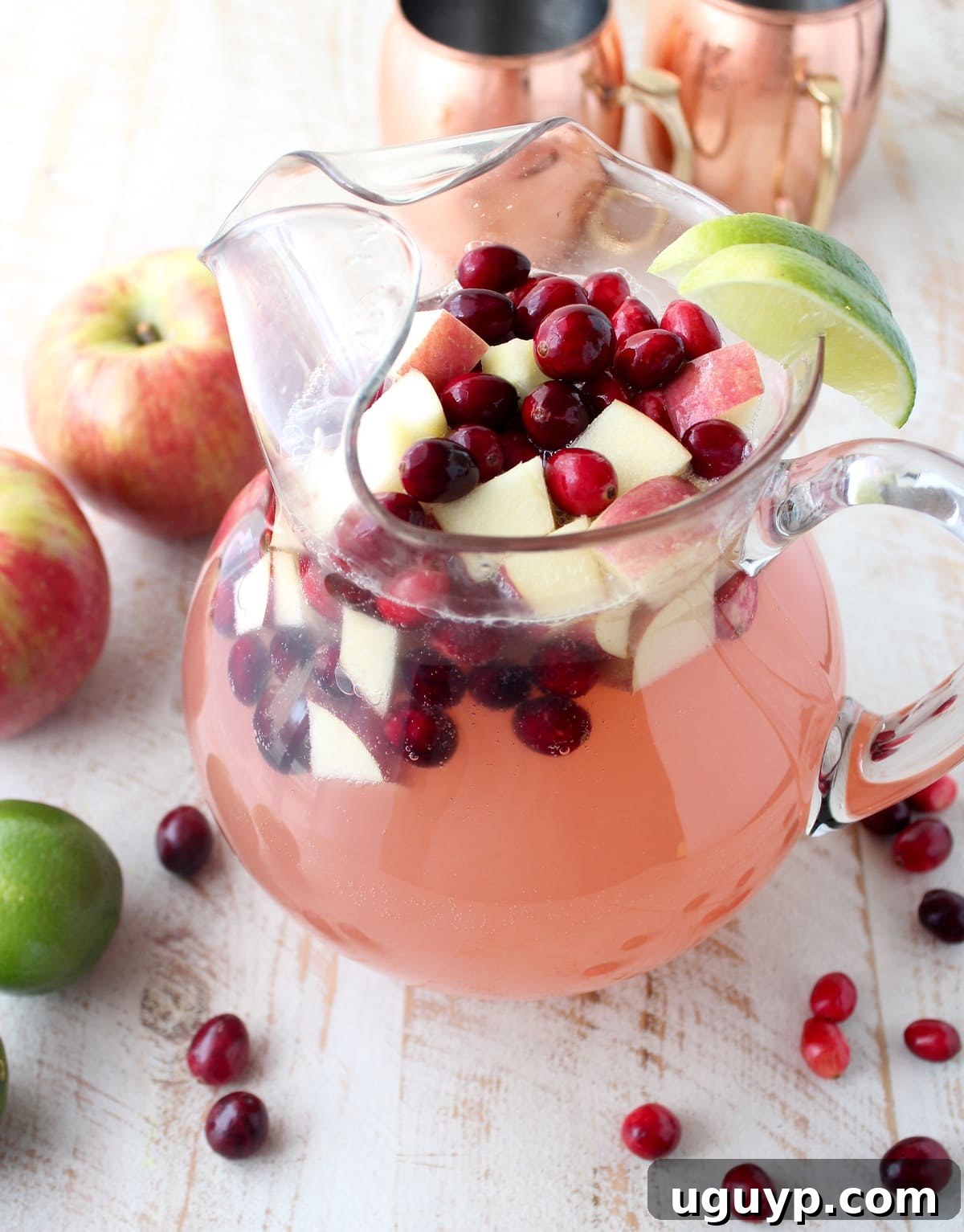 Large glass pitcher filled with cranberry apple Moscow Mule punch, garnished with fresh cranberries and diced apples, ready to be served.