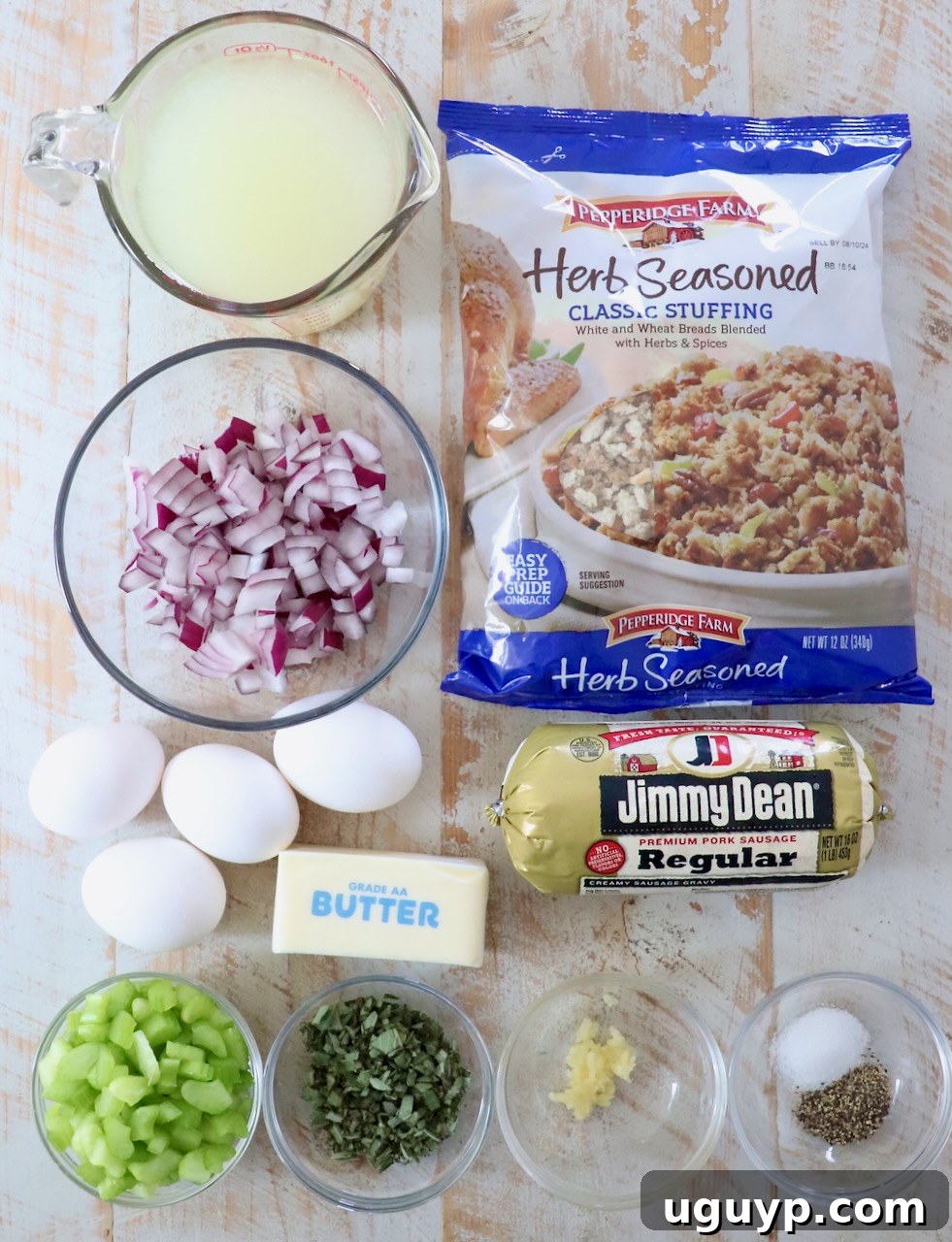A collection of fresh ingredients for making sausage stuffing, including celery, onion, sage, and a packet of sausage, arranged on a white wooden board
