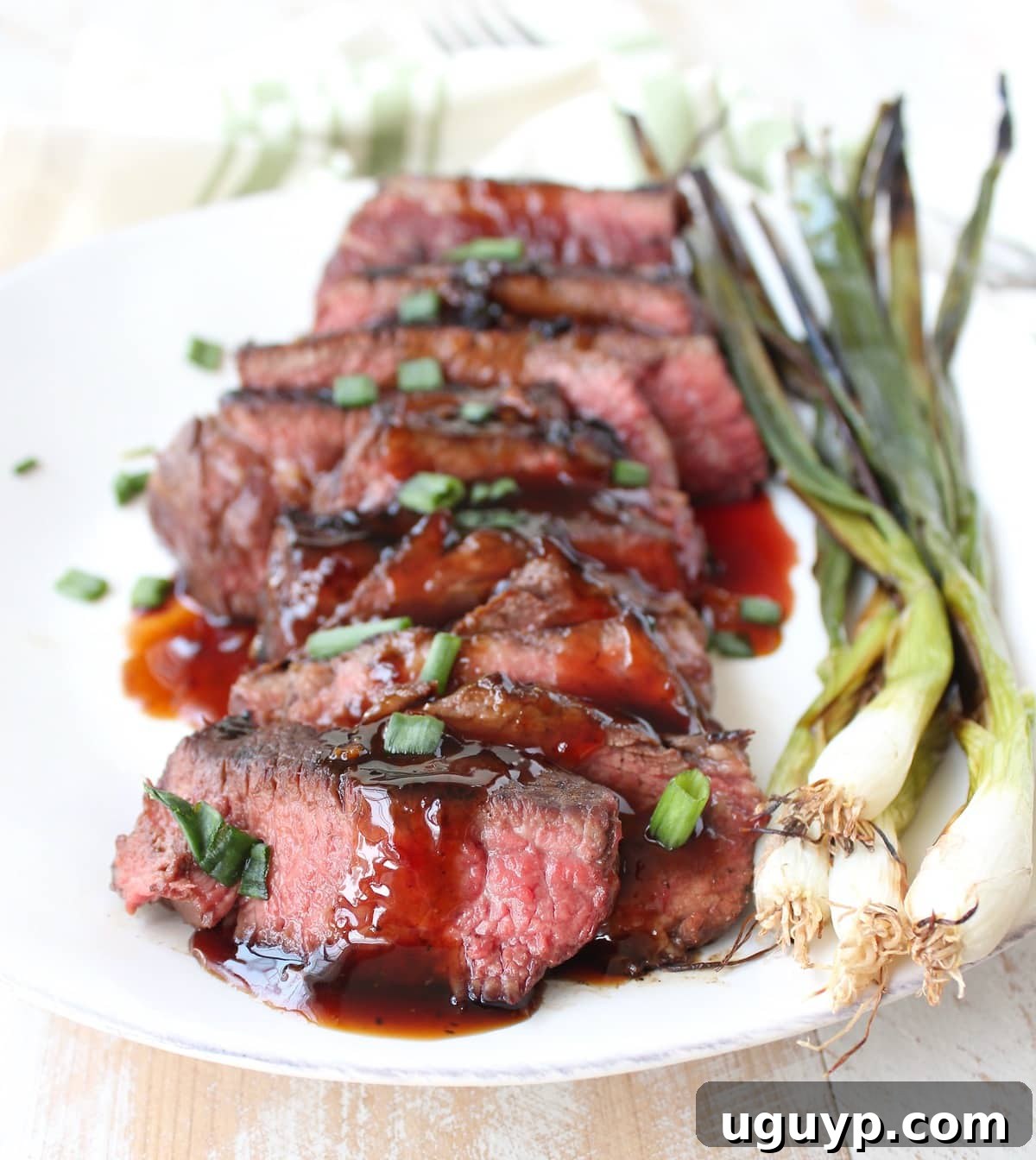 Sliced boneless beef short ribs on a plate, generously drizzled with homemade Korean BBQ sauce and garnished with fresh green onions. Grilled whole green onions are served on the side.
