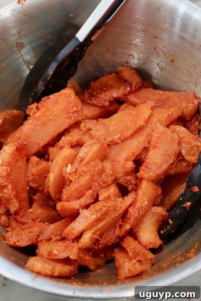 strips of raw chicken tossed in spices in metal bowl with kitchen tongs