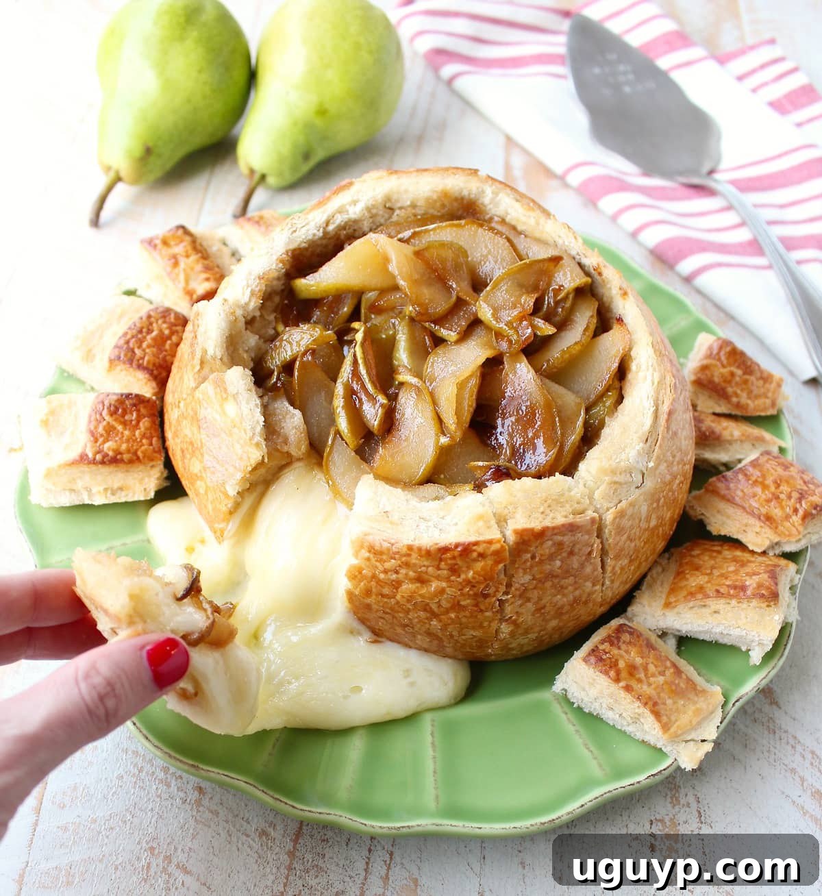 A hand reaching into a warm baked brie bread bowl to pull out a piece of bread, showcasing the rich, melted brie and caramelized pears inside.