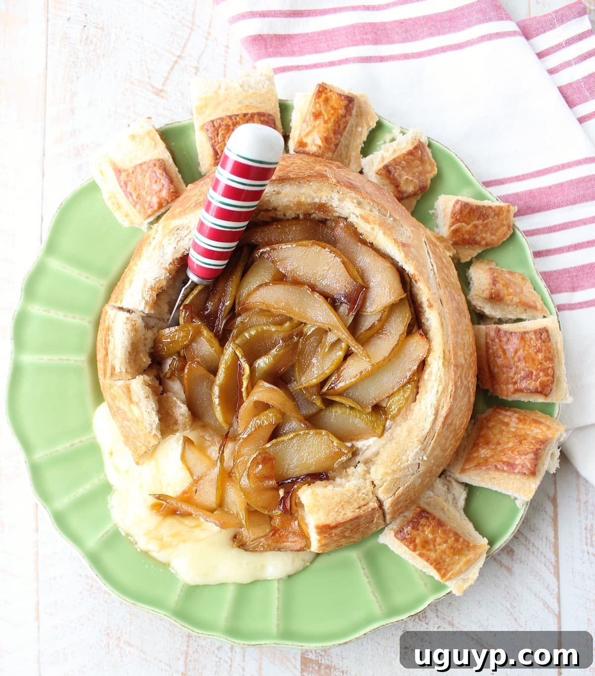 A festive holiday spread featuring the baked brie in a bread bowl with caramelized pears and honey, accompanied by a red and white cheese spreader and a decorative towel.