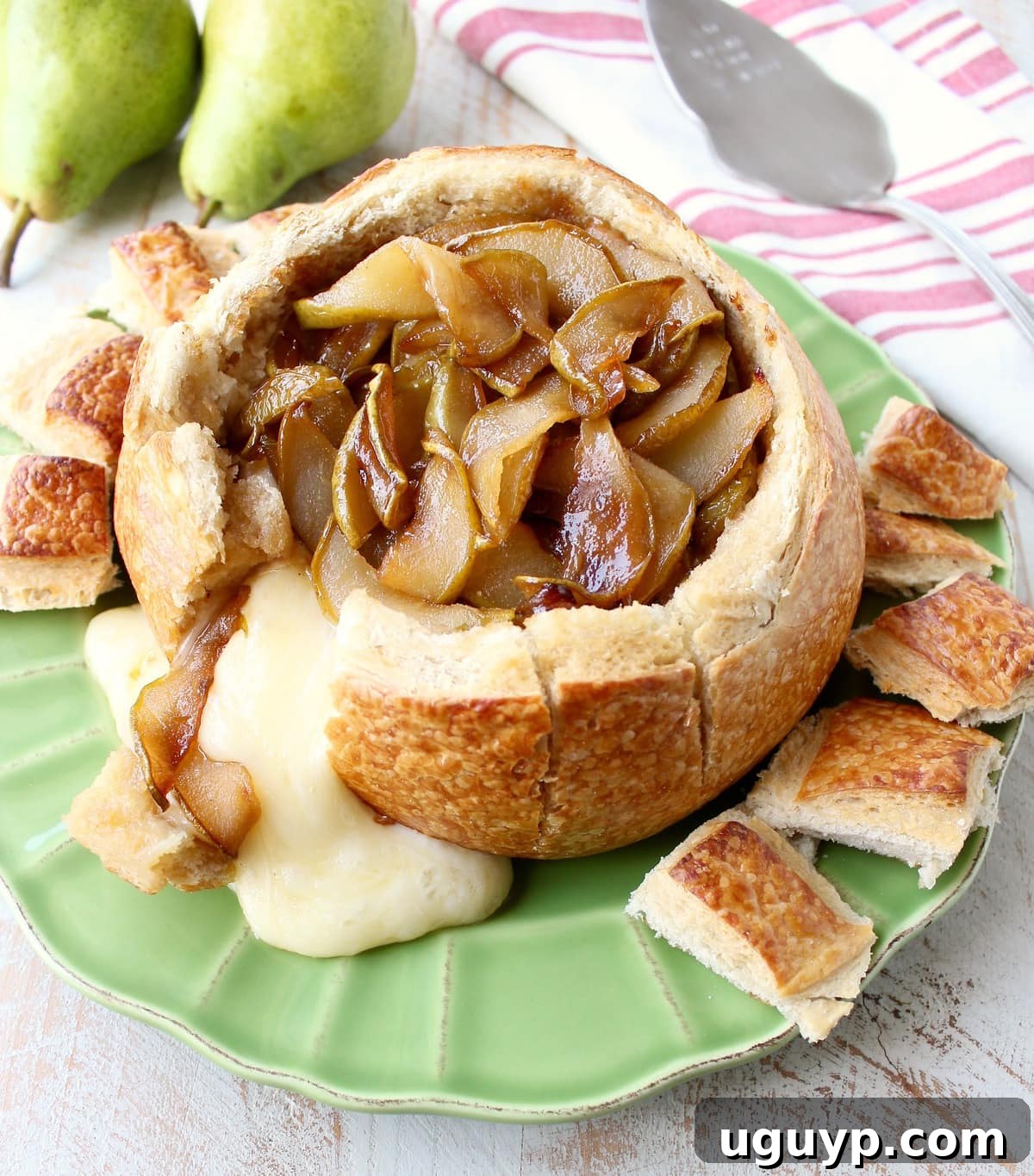 Golden baked brie elegantly overflowing from a sourdough bread bowl, adorned with sweet caramelized pears, presented on a vibrant green plate.