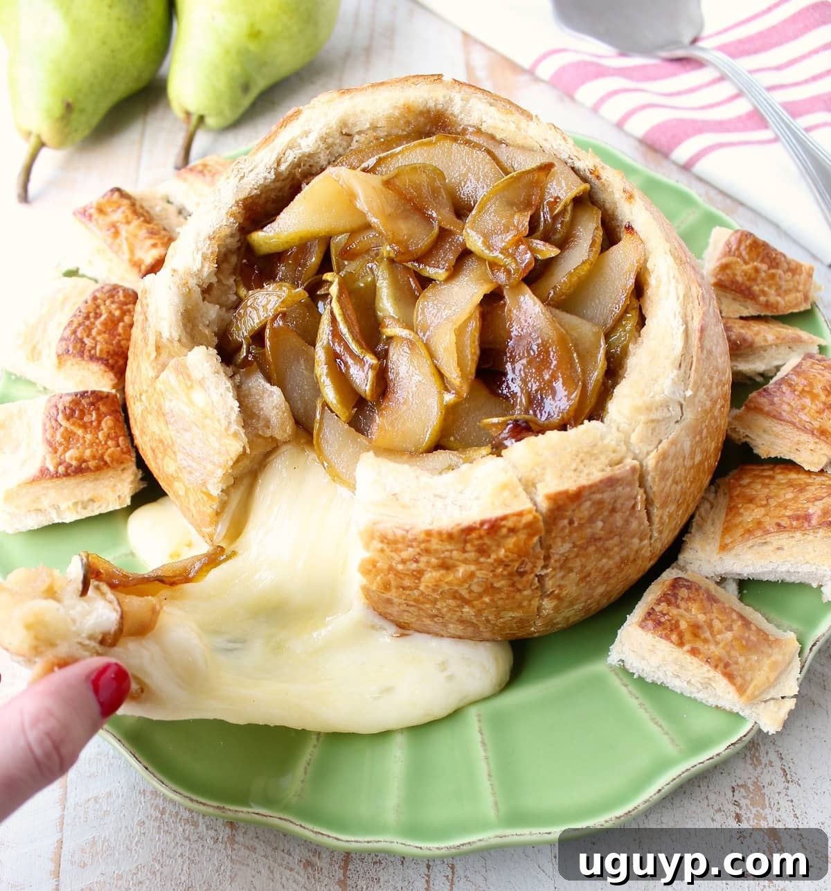 A close-up of a hand pulling a piece of warm, crusty bread from a baked brie bread bowl, revealing the gooey melted brie and caramelized pears within.