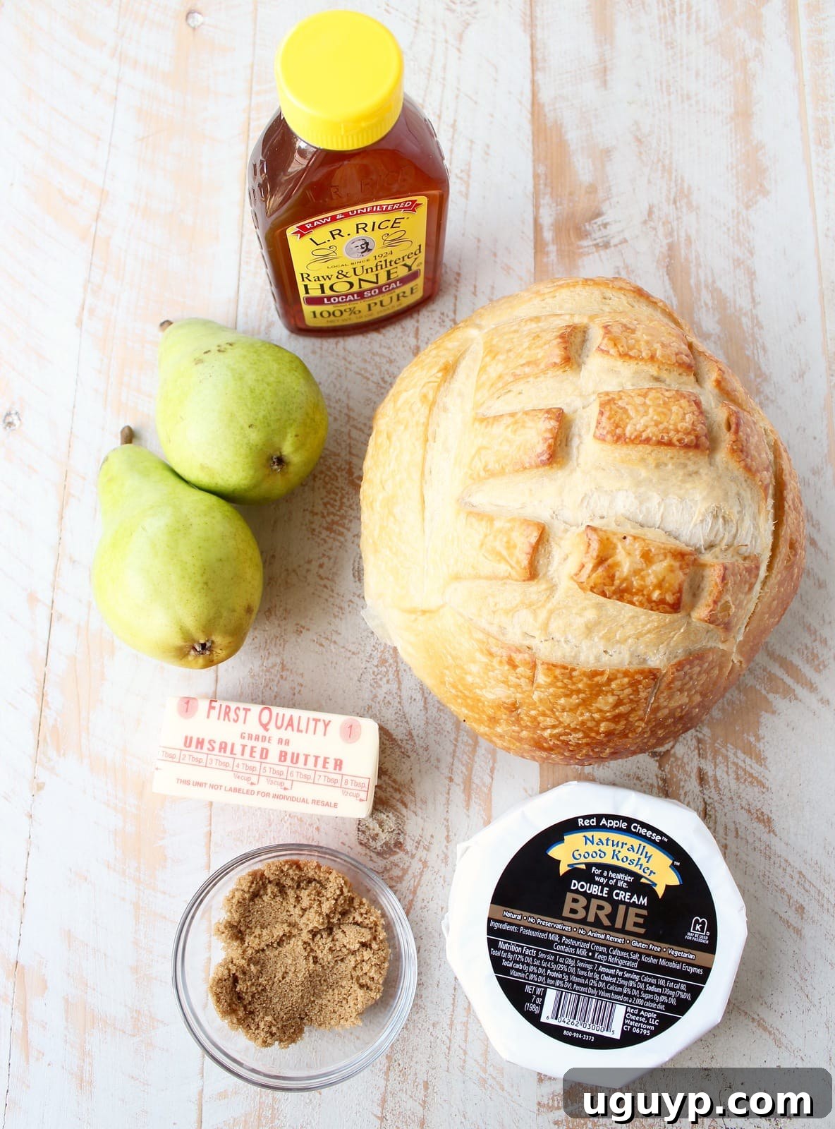 The essential ingredients for a Baked Brie Sourdough Bread Bowl: honey, fresh pears, butter, a wheel of brie, a round sourdough loaf, and brown sugar, neatly arranged.