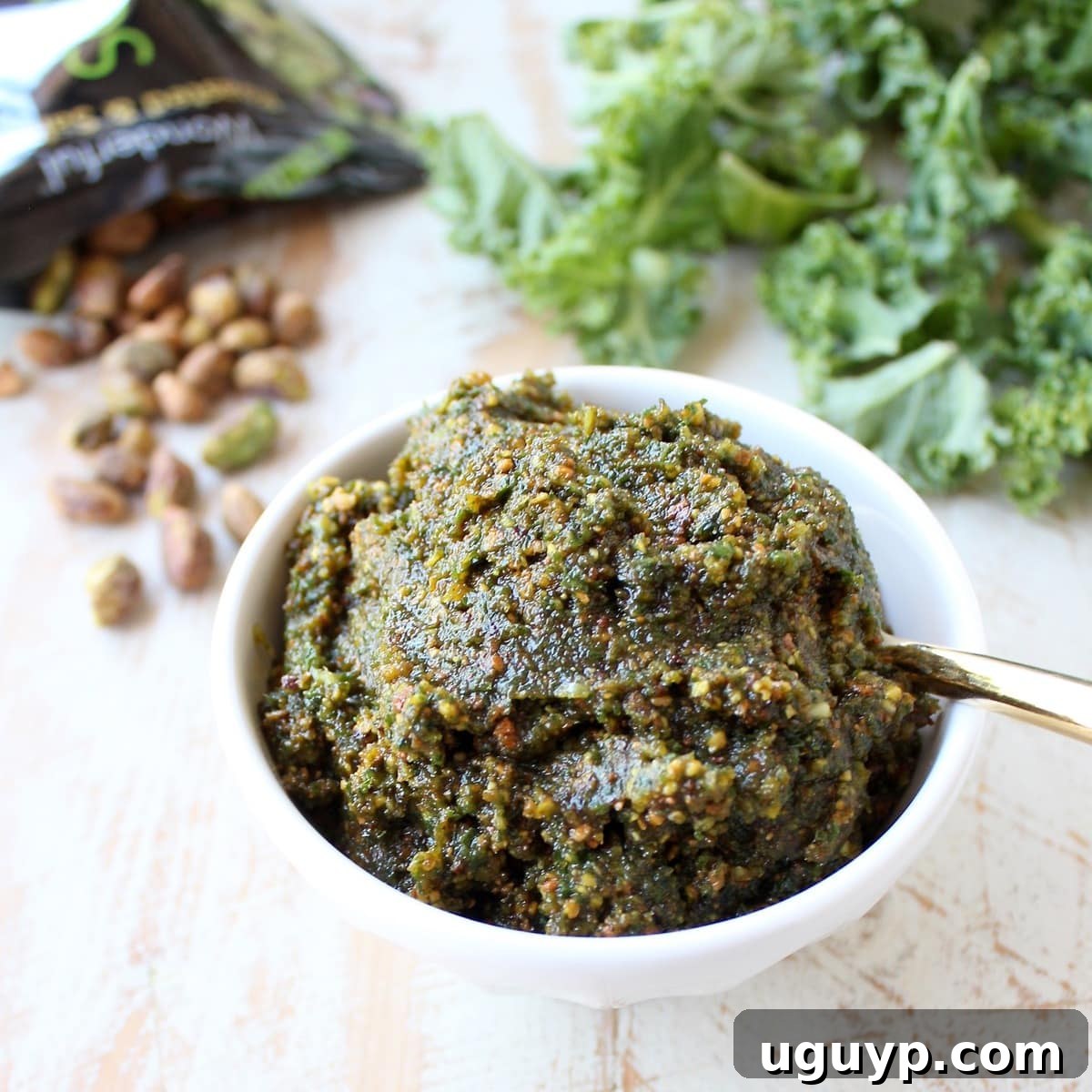 A beautifully presented bowl of vibrant green kale pistachio pesto, ready to be enjoyed. A small wooden spoon rests in the bowl, inviting you to taste.