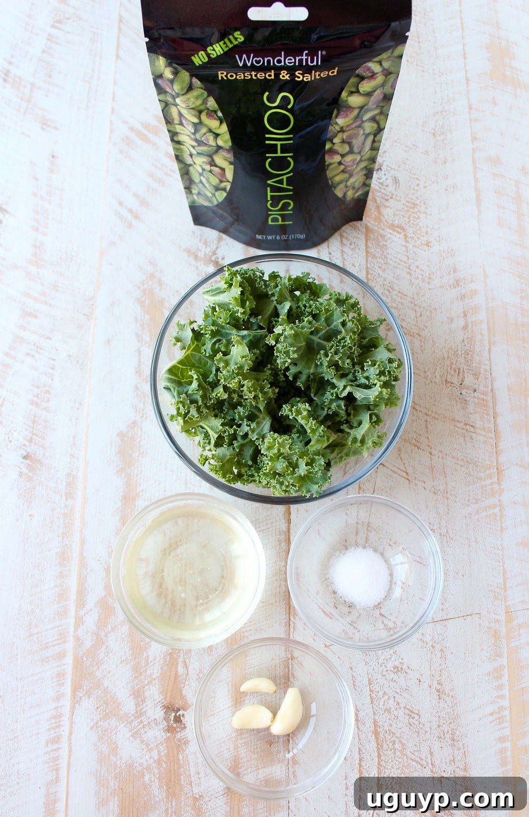 A selection of fresh ingredients for making kale pistachio pesto arranged artfully on a white wooden board, including a pile of vibrant green kale, shelled pistachios, fresh garlic cloves, and a jar of solidified coconut oil.