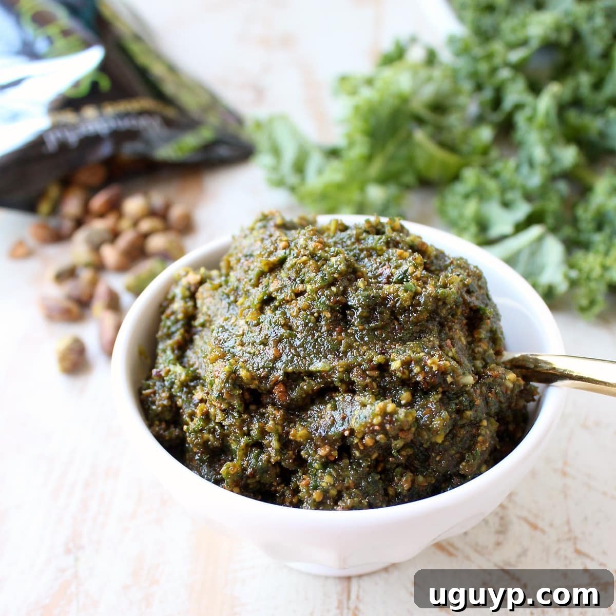 A close-up shot of freshly prepared pistachio pesto in a stylish, rustic bowl. A small wooden spoon is nestled in the pesto, and a few whole pistachios are scattered for garnish, highlighting the key ingredient.