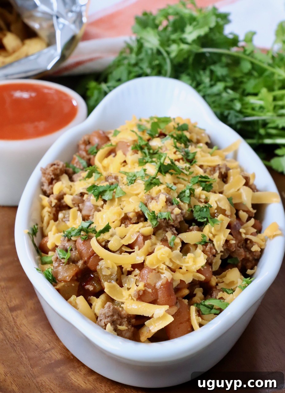 A small white dish filled with Buffalo Frito Chili Pie, generously topped with melted shredded cheddar cheese, ready to be enjoyed.