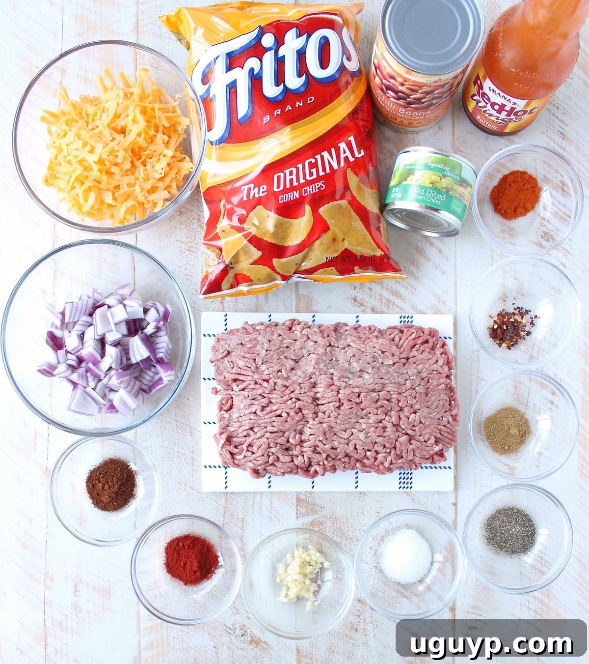 A collection of fresh ingredients for Buffalo Frito Chili Pie laid out on a rustic white wooden board, including ground beef, red onion, garlic, canned goods, spices, Fritos, and cheddar cheese.