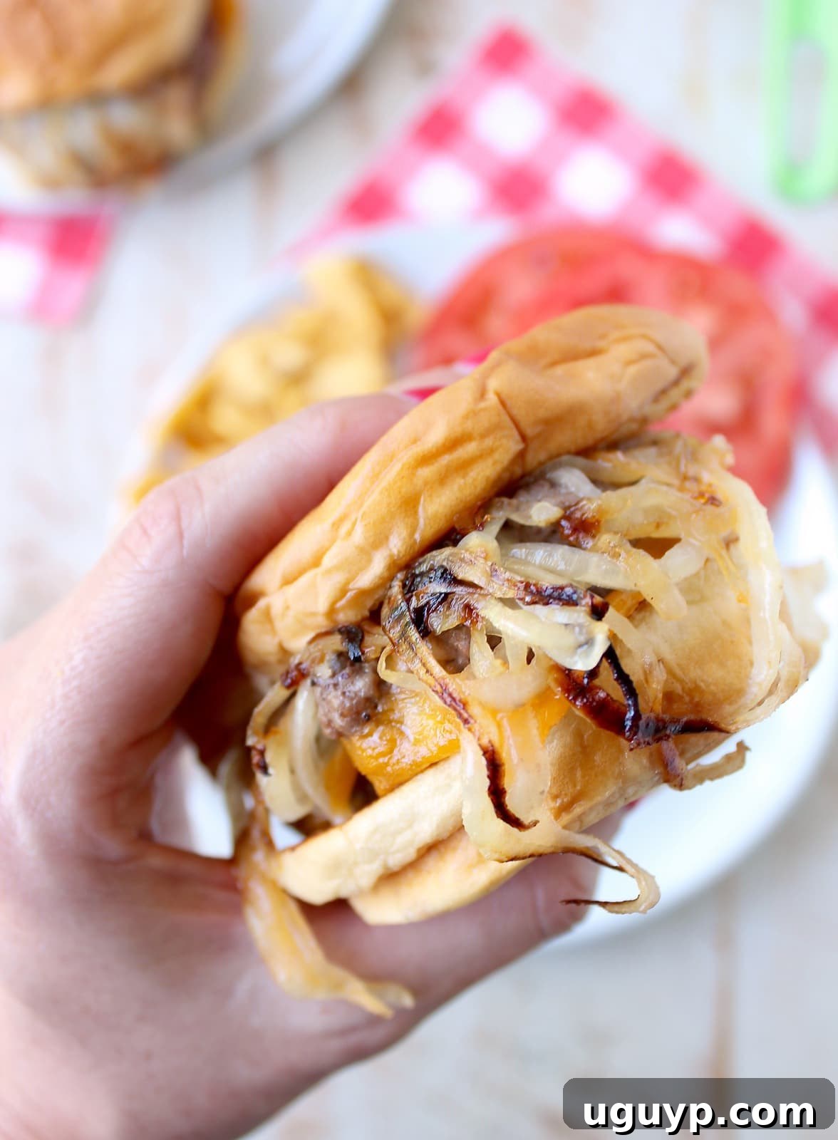A hand holding a perfectly cooked Oklahoma Fried Onion Burger, lifted from the plate to showcase its layers of beef, caramelized onions, and melted cheese.