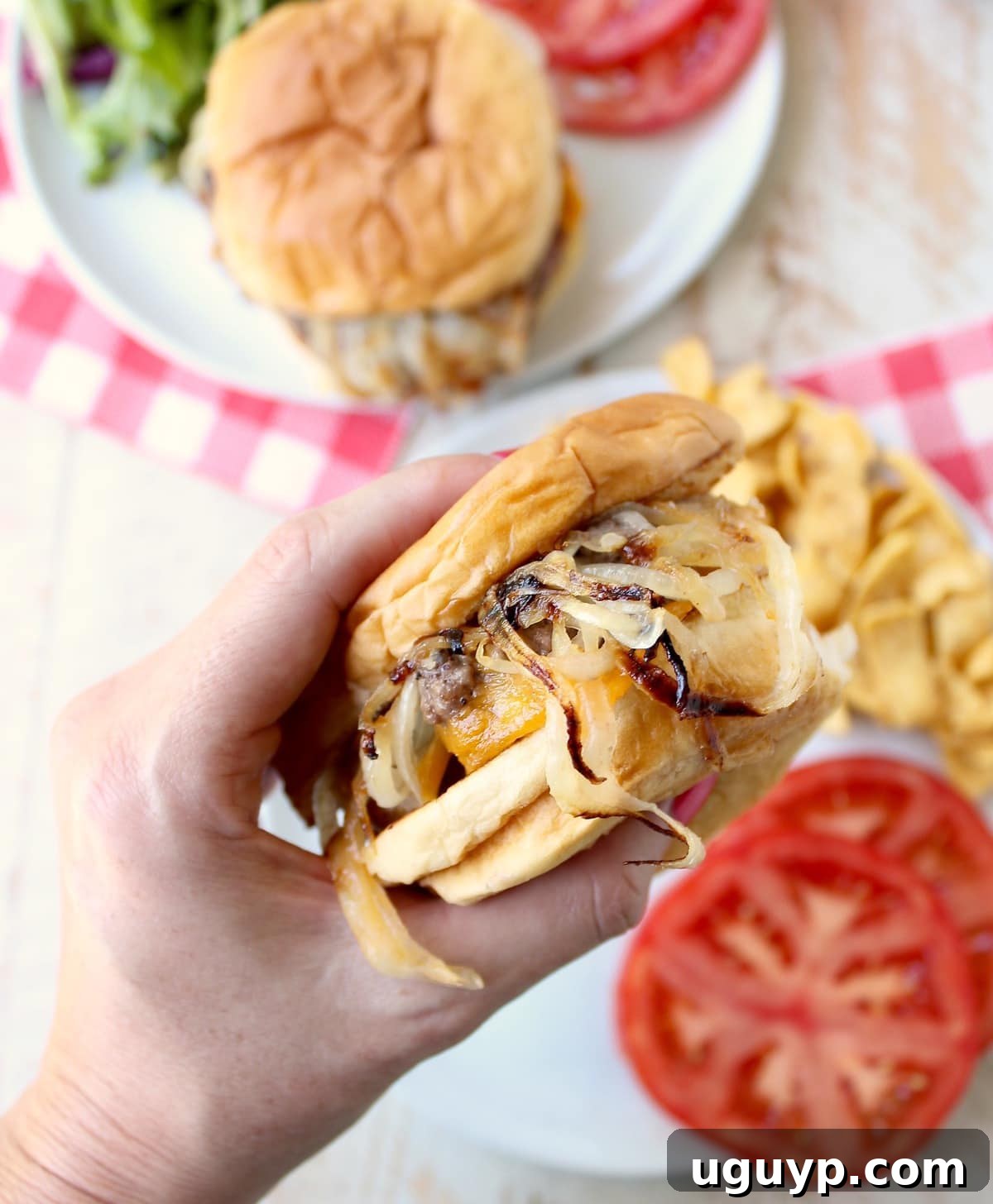A close-up shot of a hand presenting an Oklahoma Fried Onion Burger over a plate, accompanied by vibrant sliced tomatoes, highlighting its fresh ingredients.