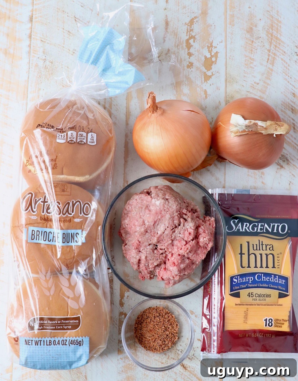 A beautiful flat lay photograph displaying all the fresh ingredients needed for Oklahoma Fried Onion Burgers on a rustic white wooden board.