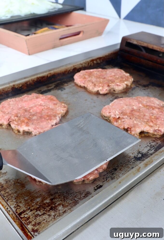 Balls of ground beef being expertly smashed into thin patties on a hot griddle with a spatula.