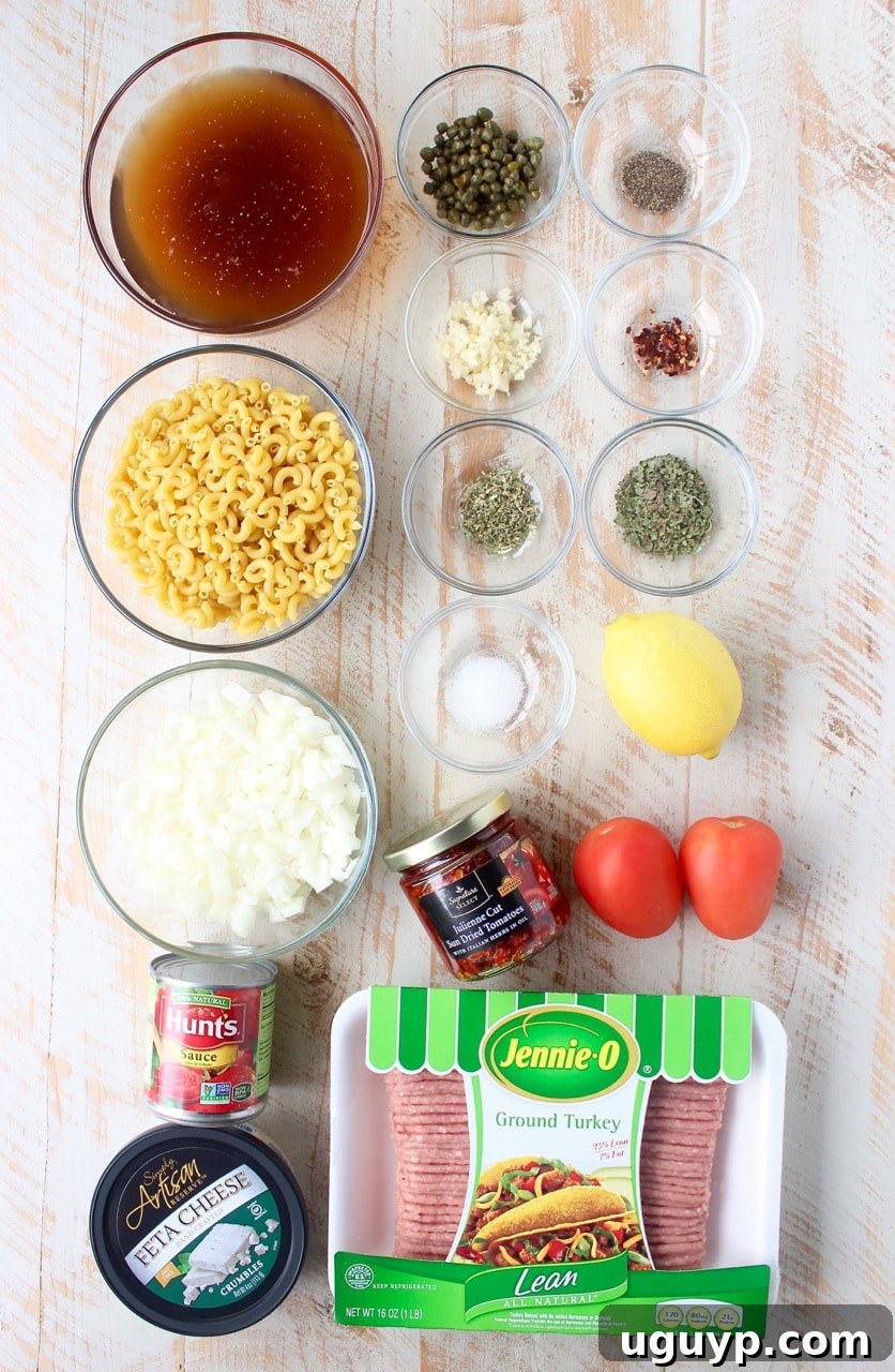 Fresh ingredients for one pot Greek pasta laid out on a rustic white wooden board, including olive oil, onions, garlic, ground turkey, various herbs, sun-dried tomatoes, Roma tomatoes, capers, tomato sauce, macaroni noodles, beef broth, lemon, and feta cheese.
