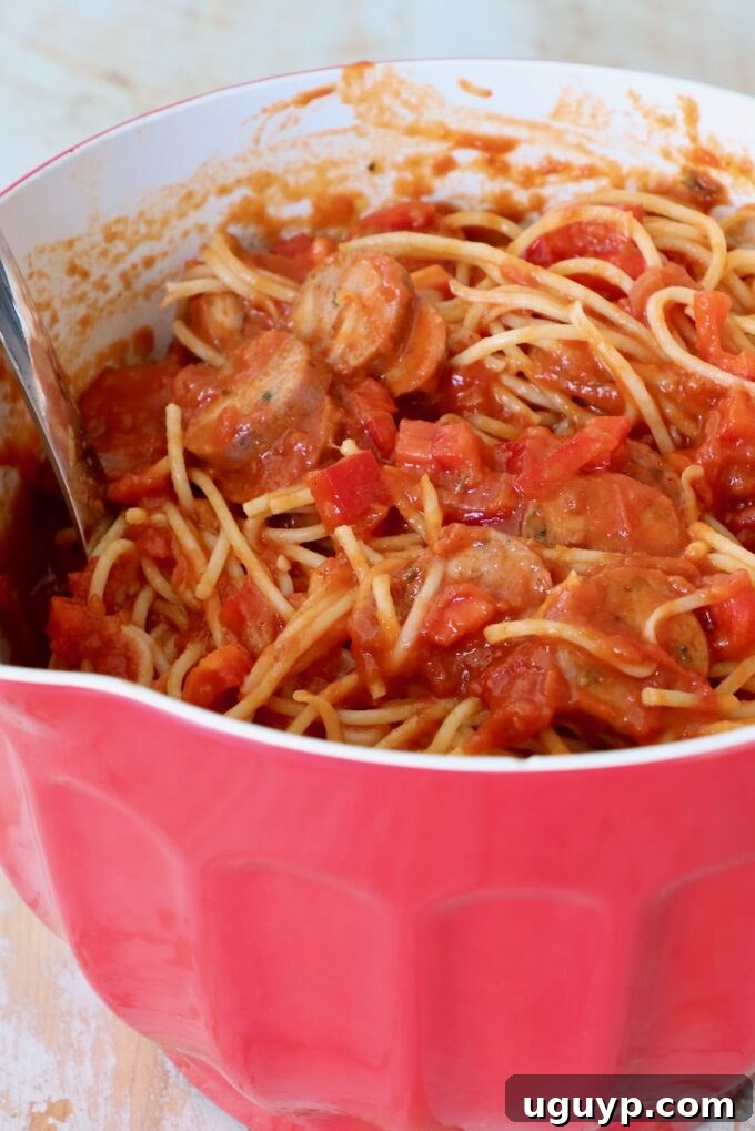 Cooked spaghetti tossed with marinara sauce, sliced sausage, and diced red bell peppers in a large mixing bowl.