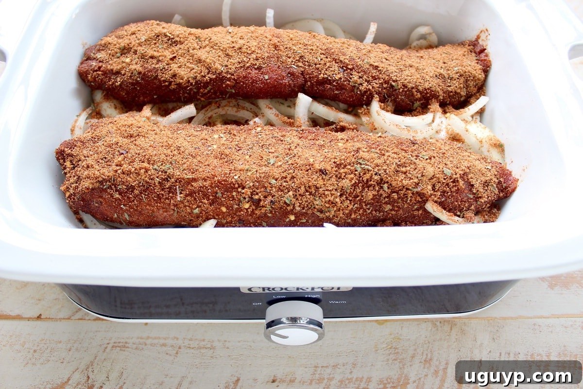 Two pork loins coated in a dry rub resting on a bed of sliced onions in a slow cooker, ready for the sauce.