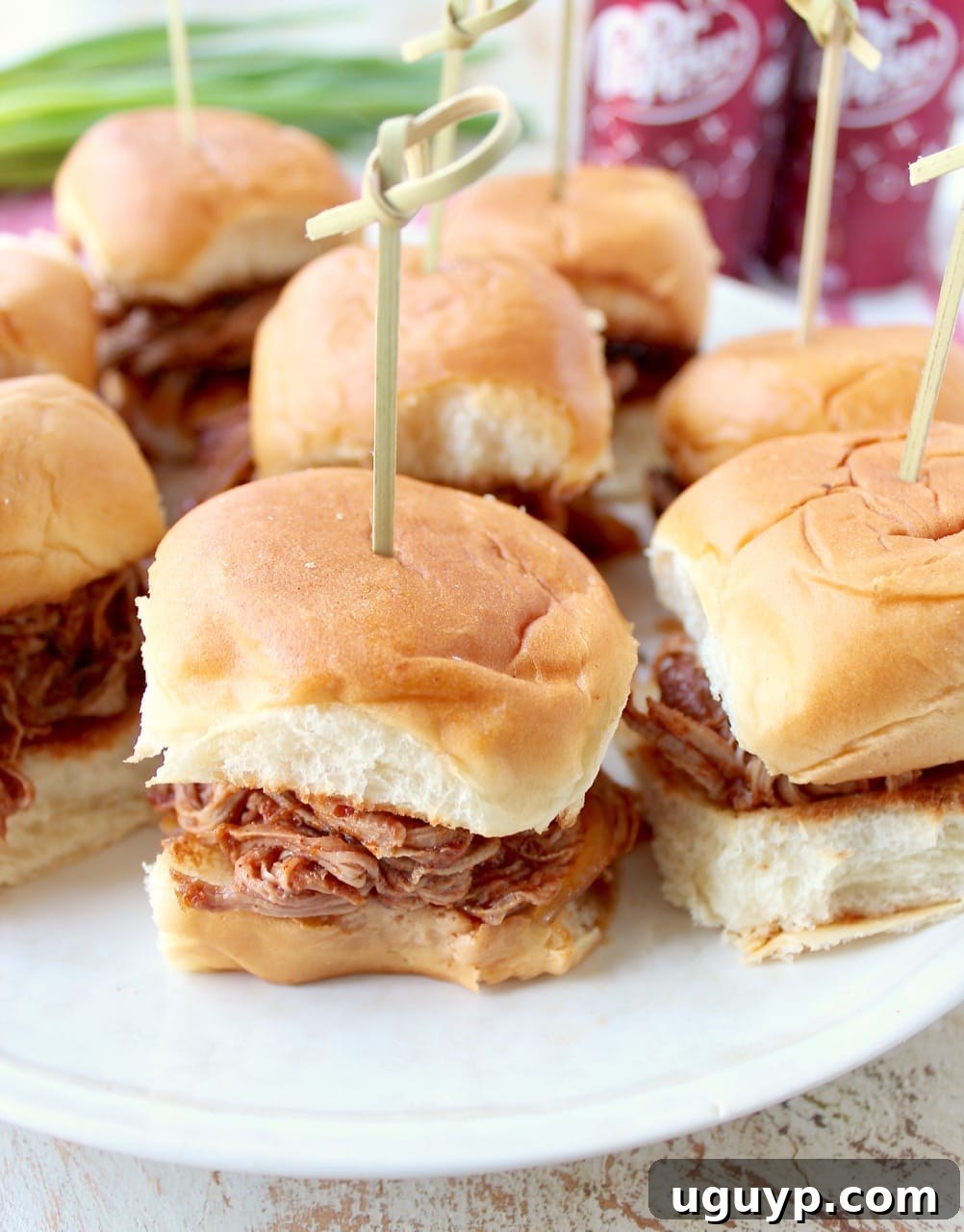 Individual Dr Pepper Pulled Pork Sliders on Hawaiian rolls, each secured with a toothpick, arranged on a white serving plate.
