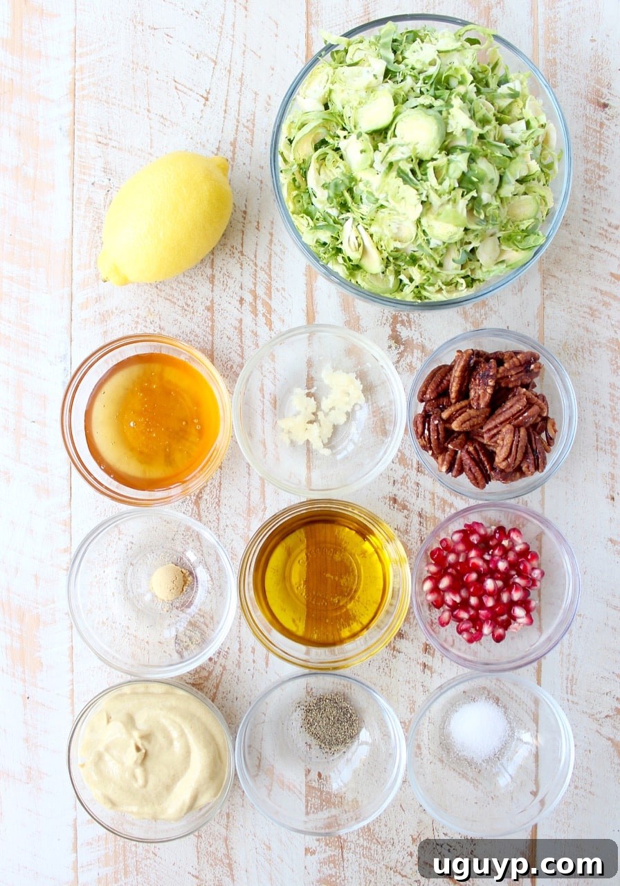 Fresh ingredients for a Brussels sprout salad laid out on a white wooden board, including whole Brussels sprouts, a honeycrisp apple, a pomegranate, and a small bowl of honey dijon dressing, ready for preparation.